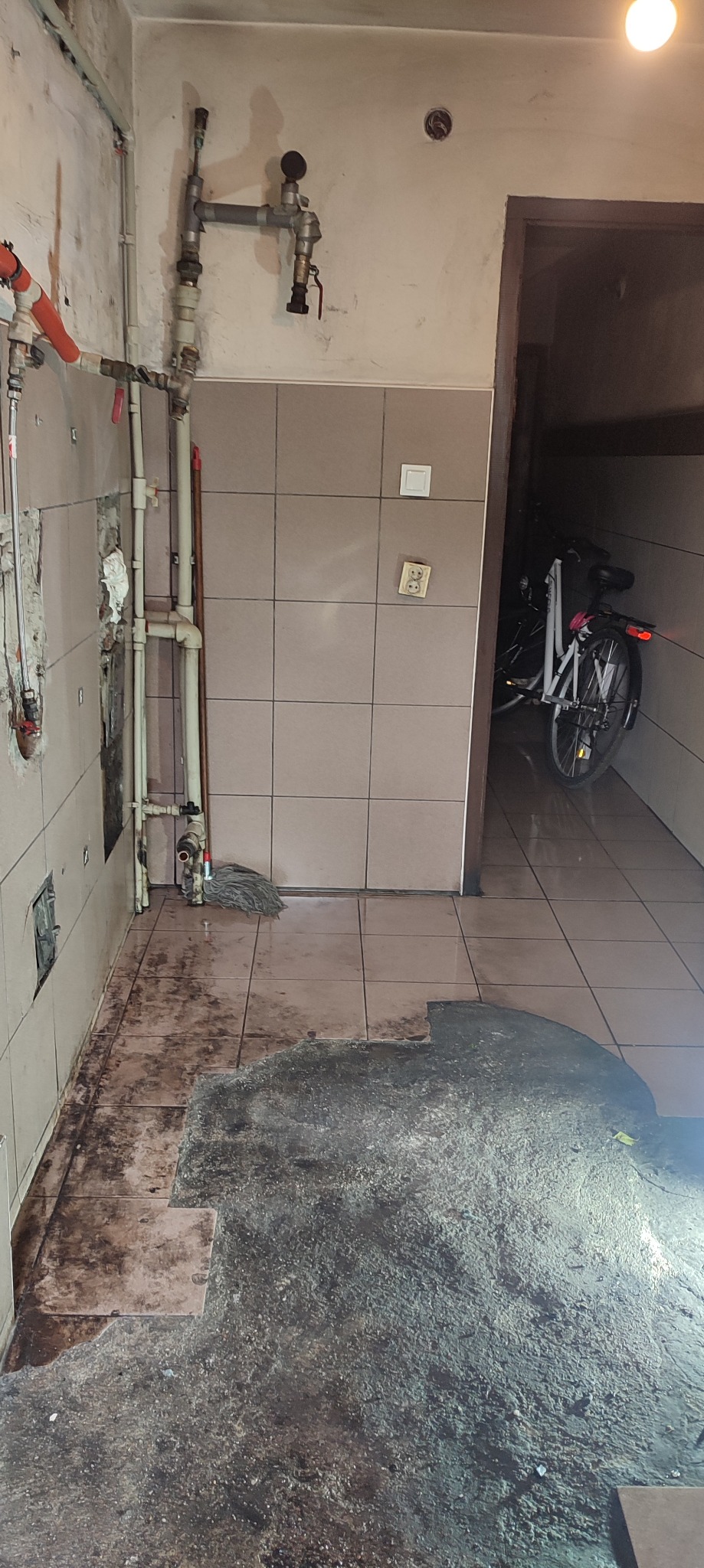 Podłoga po remoncie z widocznymi śladami kleju, resztkami płytek i mopem przy rurach, rower w tle.