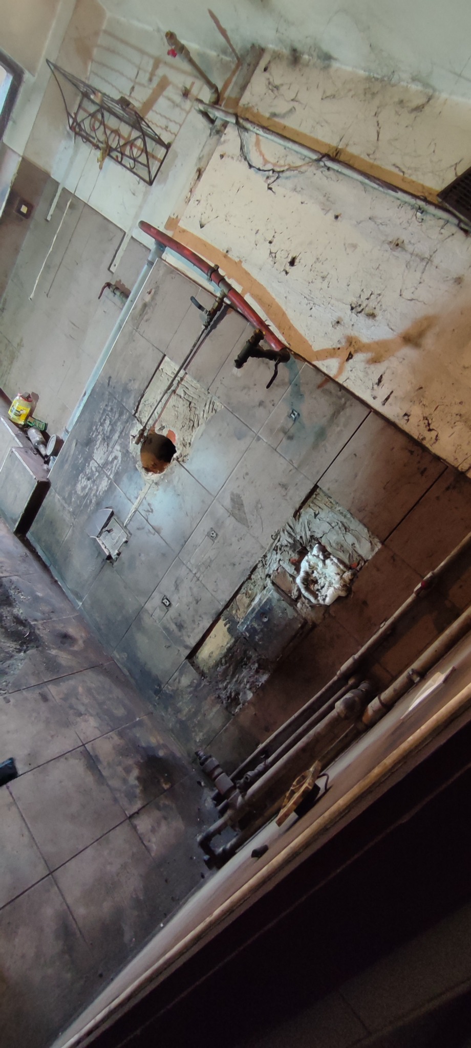 Zniszczona ściana z odchodzącymi płytkami, widoczne rury, instalacje i otwór w ścianie. Na podłodze pozostałości po remoncie i zabrudzenia.
