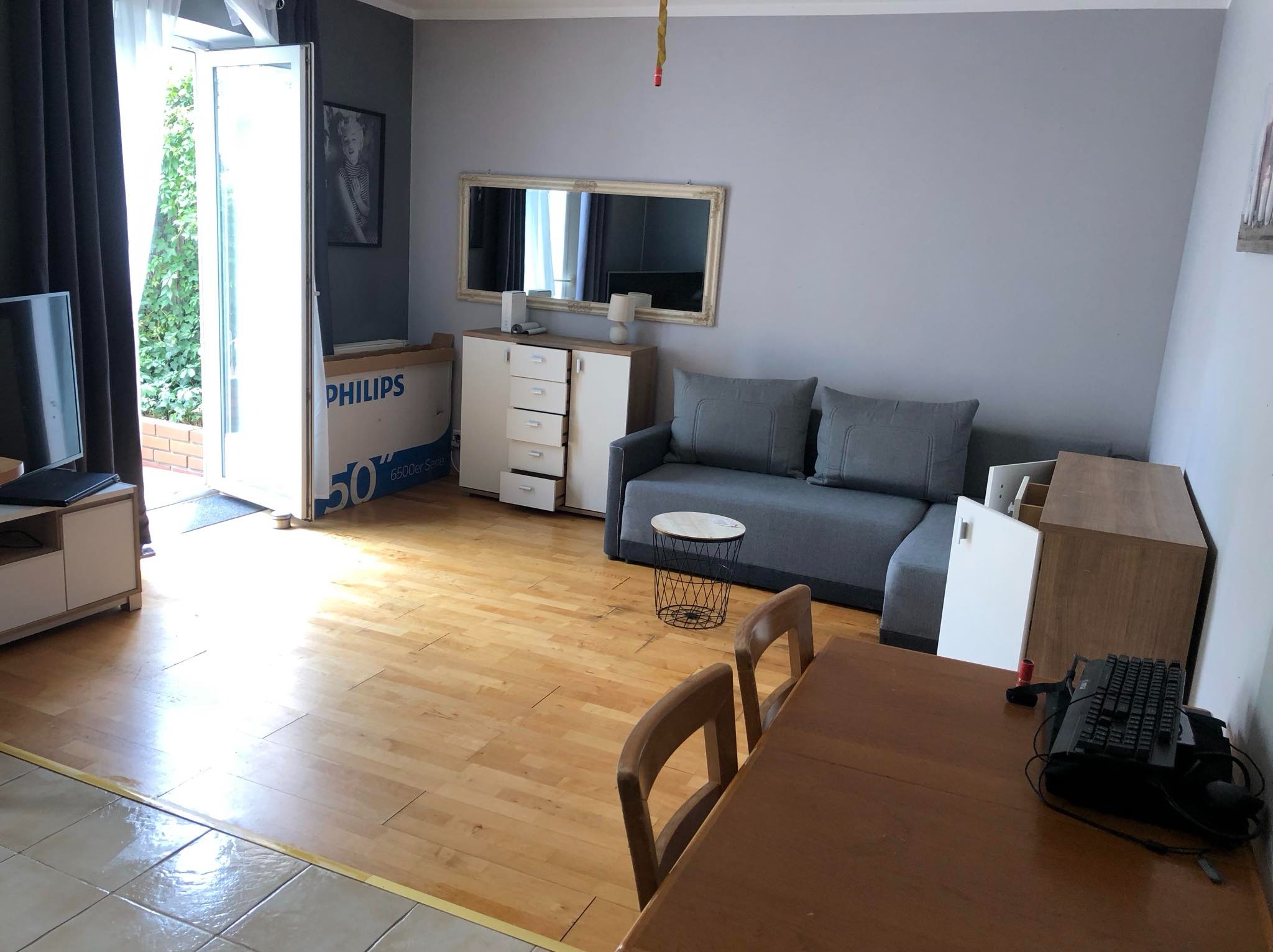 Widok na salon z drewnianą podłogą, szarą sofą, stolikiem kawowym z metalową podstawą, otwartym balkonem i widocznym kartonem po telewizorze Philips.