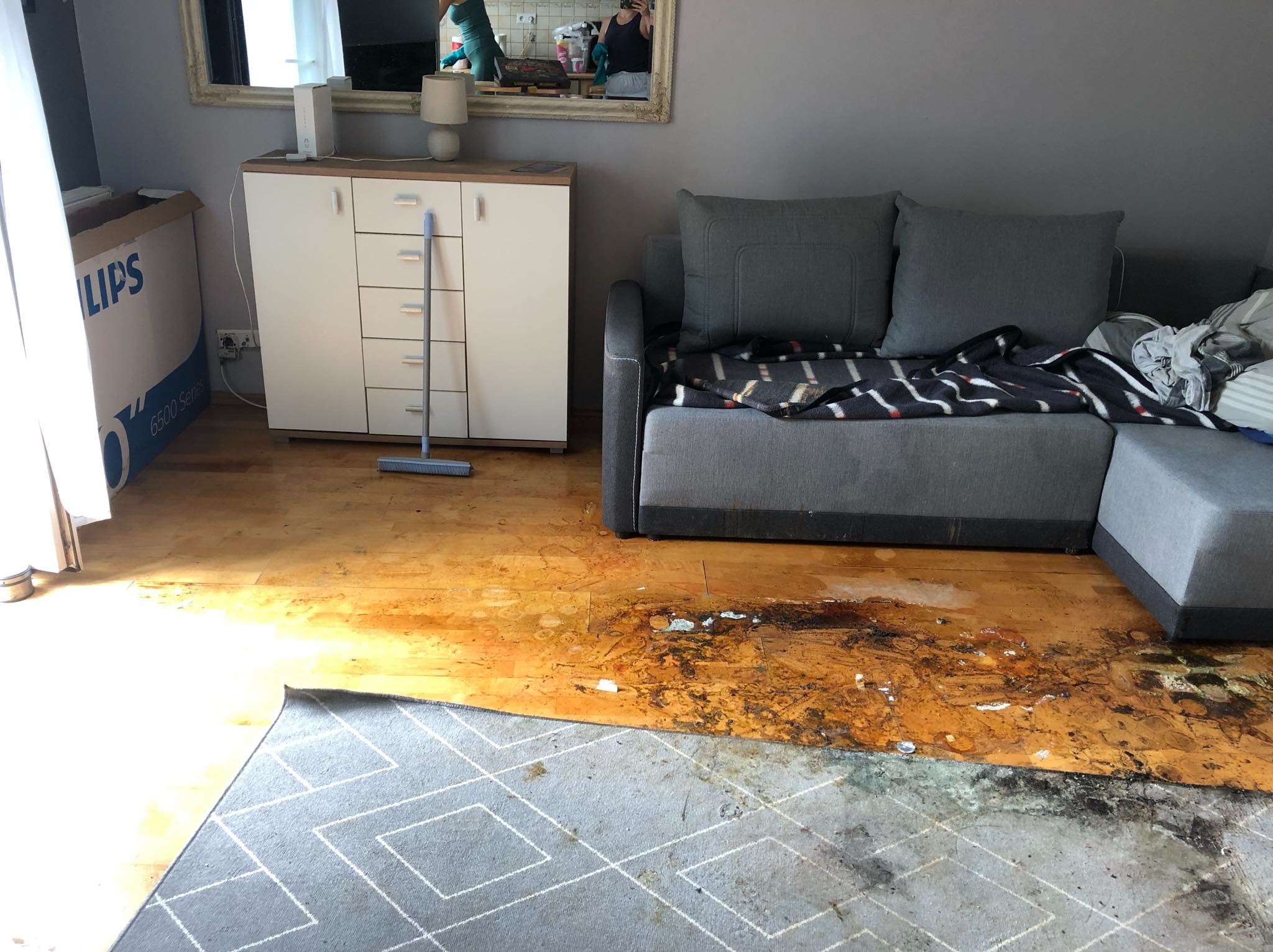 Pokój po zalaniu: zniszczona podłoga drewniana, plamy na dywanie w geometryczne wzory, szary narożnik z kocem w paski, biała komoda z lustrem, na którym widać odbicie osoby robiącej zdjęcie.