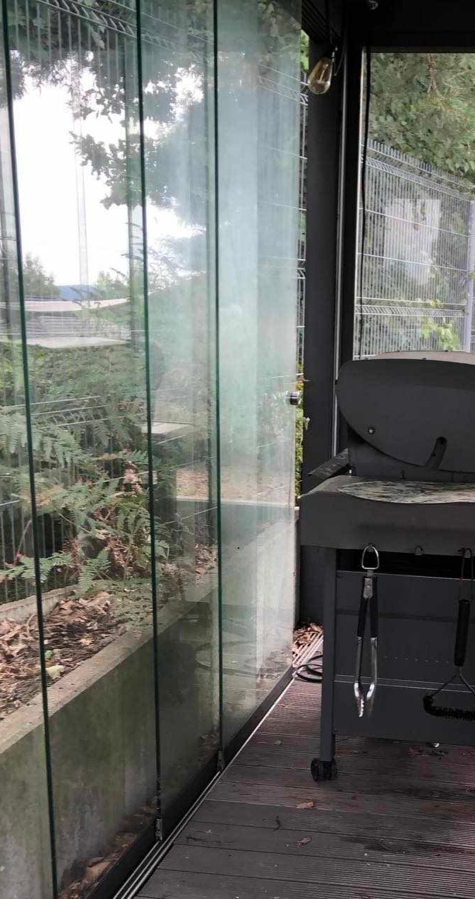 Zabrudzone, szklane panele przesuwne tarasu z widocznymi smugami i zaciekami, obok grill na kółkach z akcesoriami, widok z zewnątrz.
