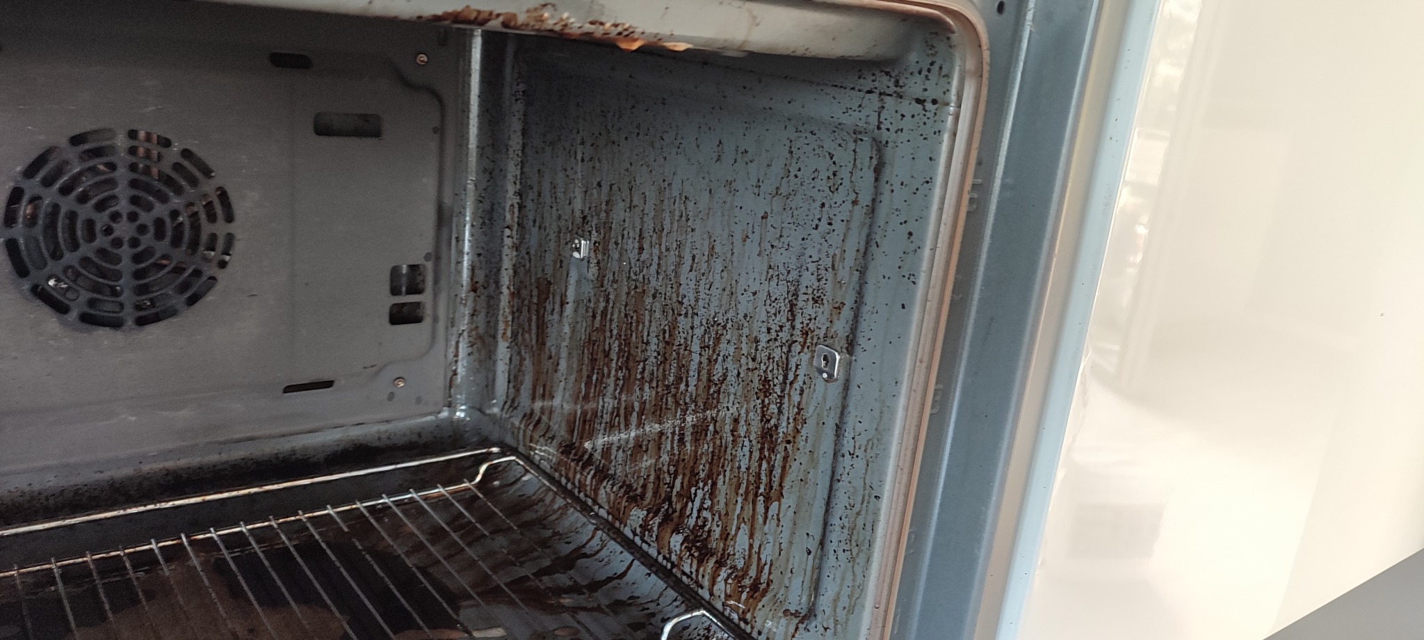 Wnętrze brudnego piekarnika z przypalonym tłuszczem i resztkami jedzenia na ściankach, kratce i wentylatorze, widok 'przed' czyszczeniem.