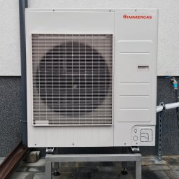 Solo24.PL - Zewnętrzna jednostka pompy ciepła marki Immergas, zamontowana na metalowej konstrukcji z regulowanymi nóżkami, przy ścianie budynku z widocznym oknem i rurą spustową.