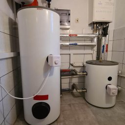 Solo24.PL - Dwa białe zasobniki ciepłej wody użytkowej (CWU) marki Drazice i A.J., z widocznymi przewodami instalacji CO i CWU, zamontowane w pomieszczeniu z wykończeniem w płytkach ceramicznych.