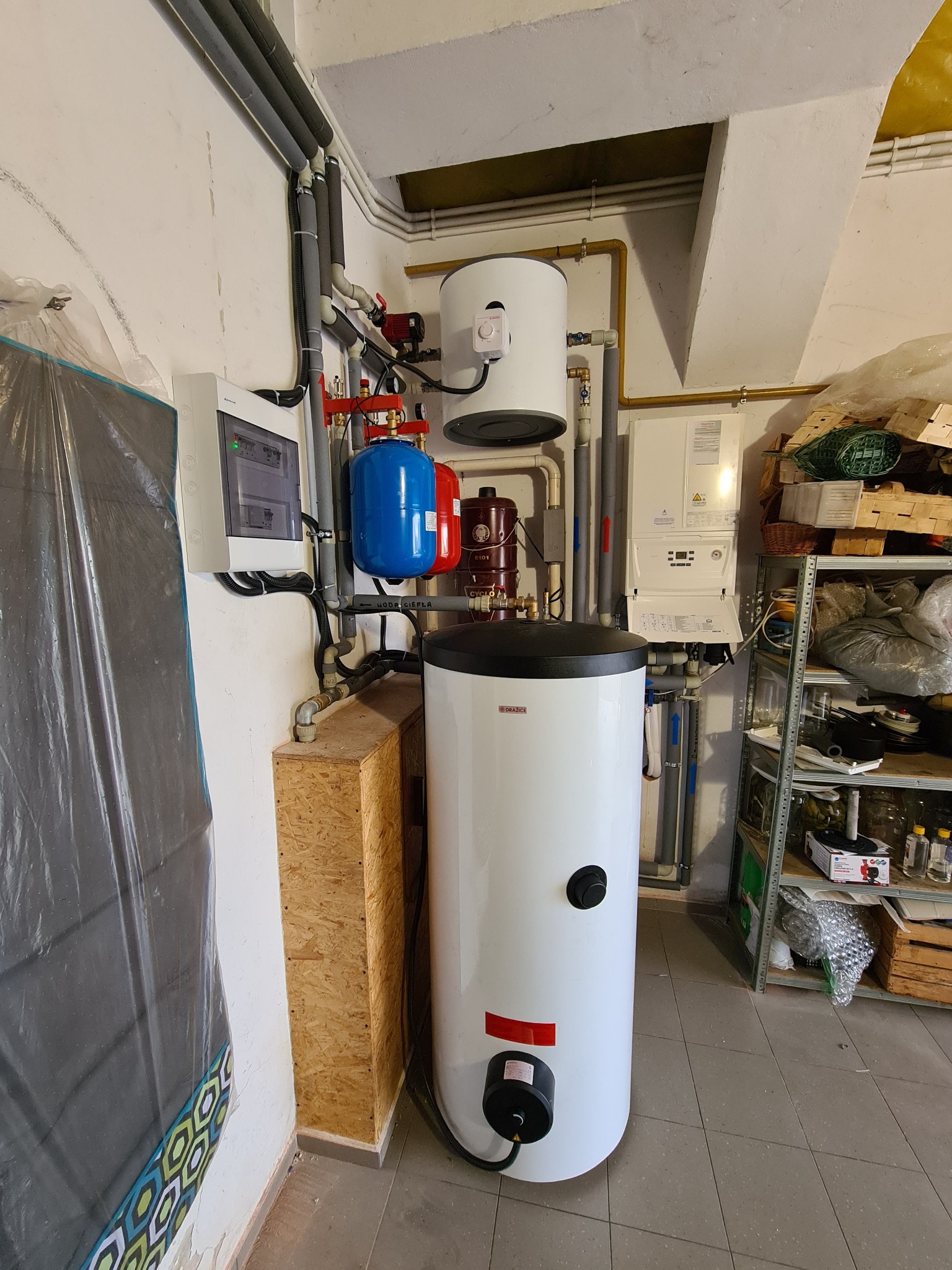 Bojler elektryczny z elementami instalacji hydrauliczej, w tle widoczna instalacja gazowa, zabezpieczenia oraz metalowy regał z różnymi przedmiotami.