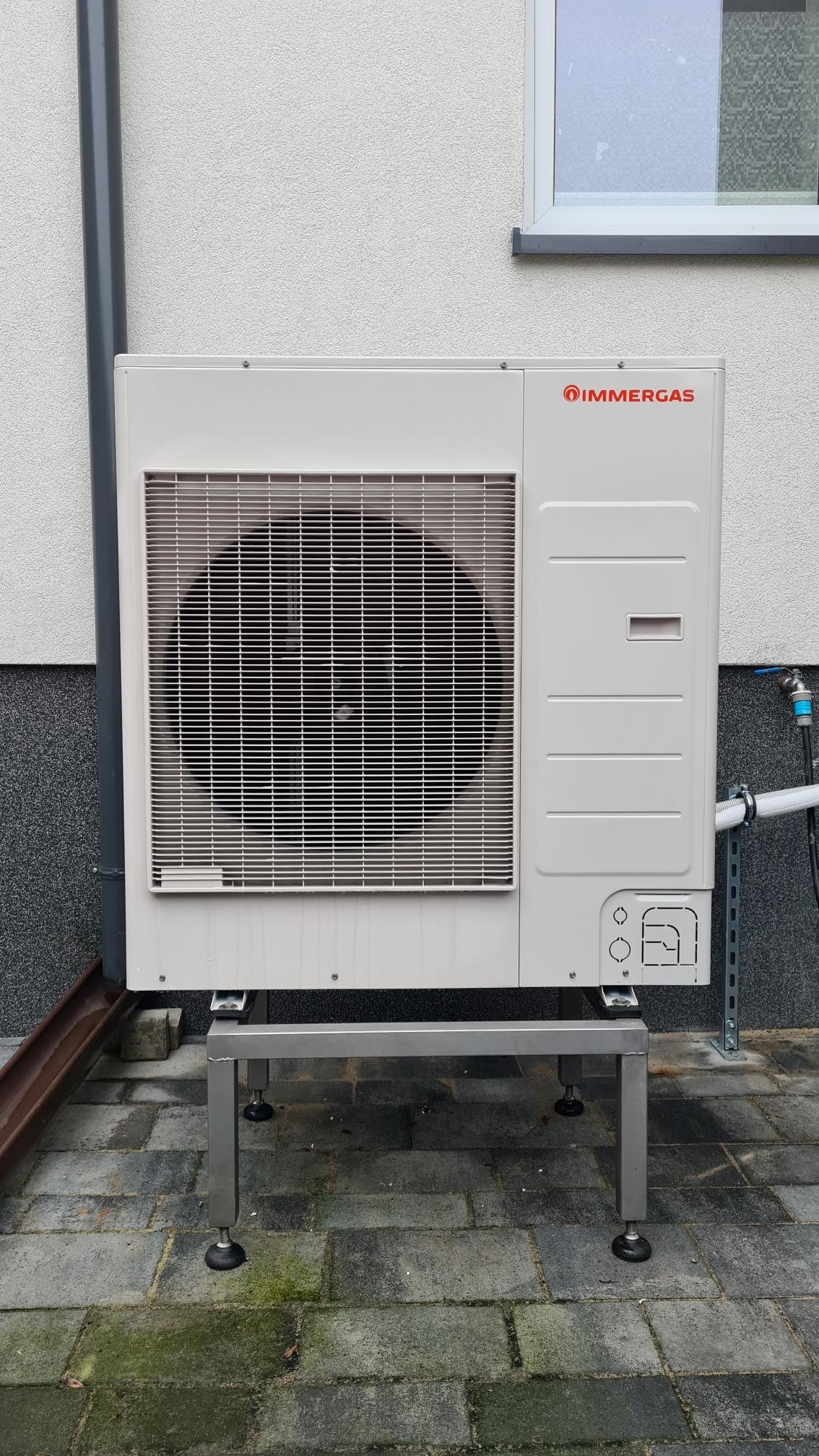 Zewnętrzna jednostka pompy ciepła marki Immergas, zamontowana na metalowej konstrukcji z regulowanymi nóżkami, przy ścianie budynku z widocznym oknem i rurą spustową.
