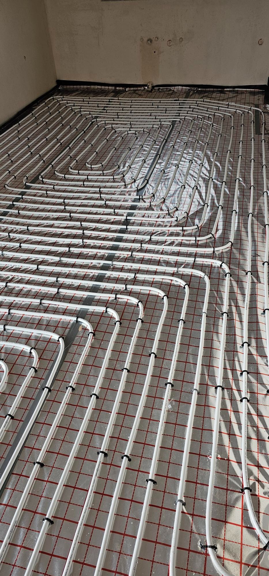 Białe rury ogrzewania podłogowego, mocowane czarnymi uchwytami do siatki z czerwonymi liniami na folii izolacyjnej, przygotowane do zalania wylewką.