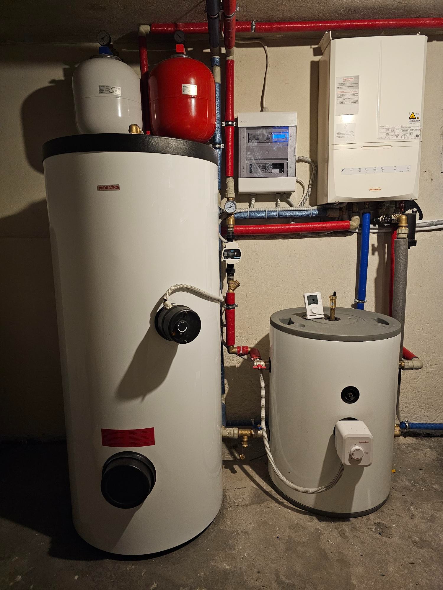 Dwa białe zasobniki ciepłej wody użytkowej marki Drazice, jeden większy z czarnym elementem regulacyjnym, drugi mniejszy z białym termostatem, instalacja CO z czerwonymi rurami, wiszący piec gazowy...