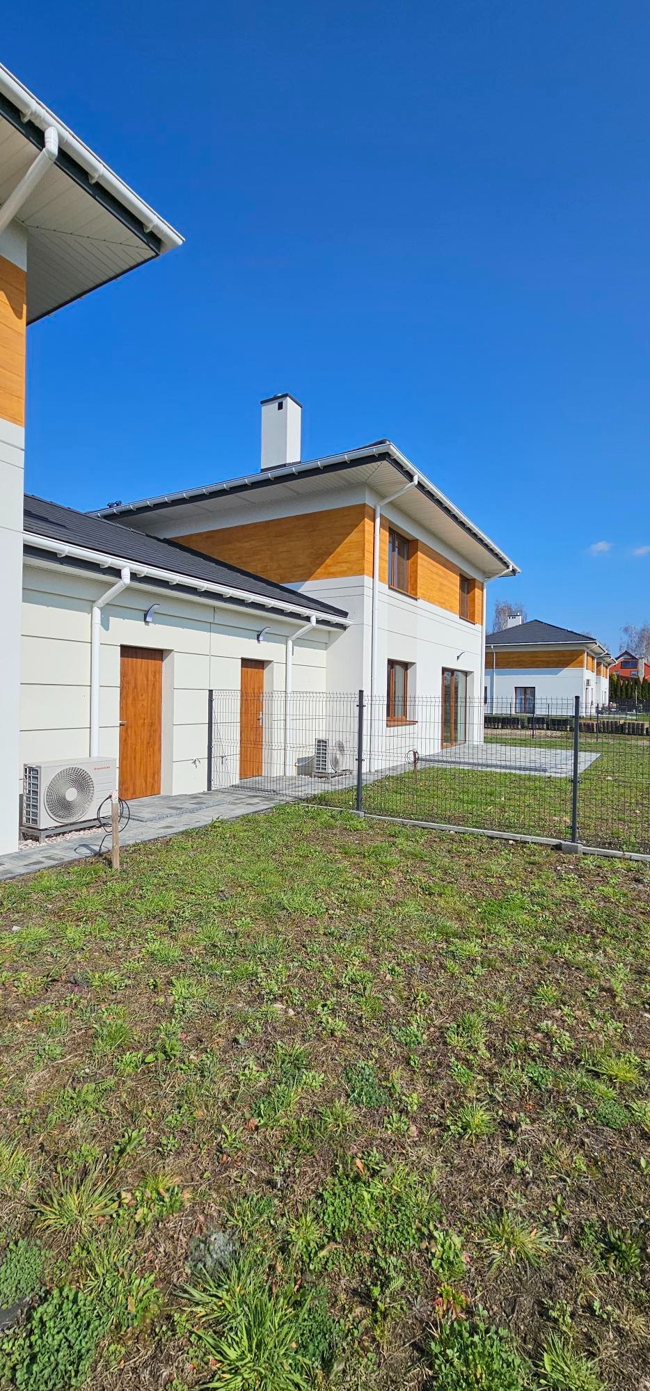 Nowoczesny dom jednorodzinny z klimatyzatorem zewnętrznym zamontowanym na ścianie, widoczny fragment ogrodzenia panelowego i trawnik.