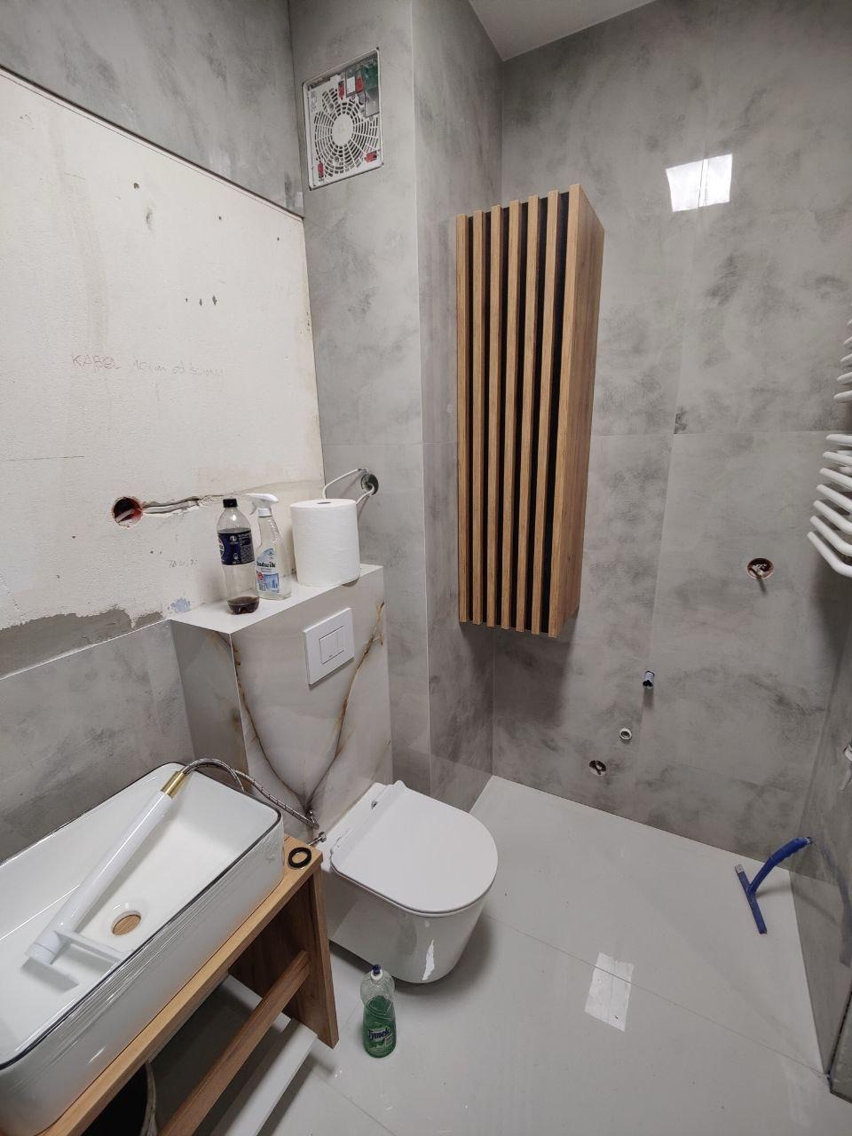 Szara łazienka w trakcie wykańczania: biała misa WC, umywalka na drewnianym stelażu, wisząca szafka z lamelami, wentylator. Na ścianach imitacja marmuru.