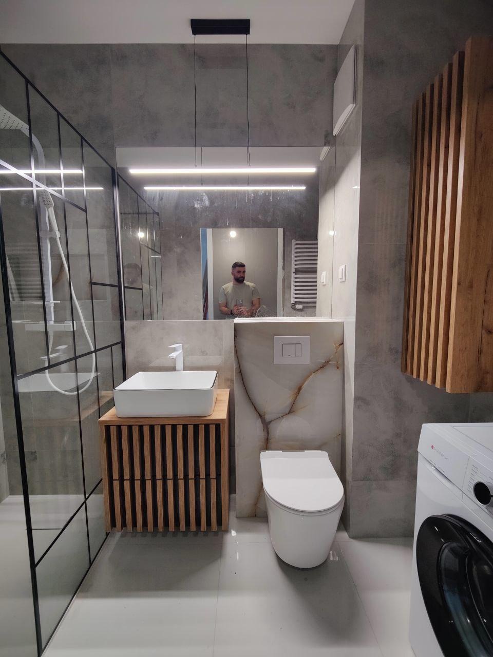 Nowoczesna łazienka z kabiną prysznicową typu walk-in, umywalką na drewnianej szafce i podwieszaną toaletą. W lustrze odbija się osoba.