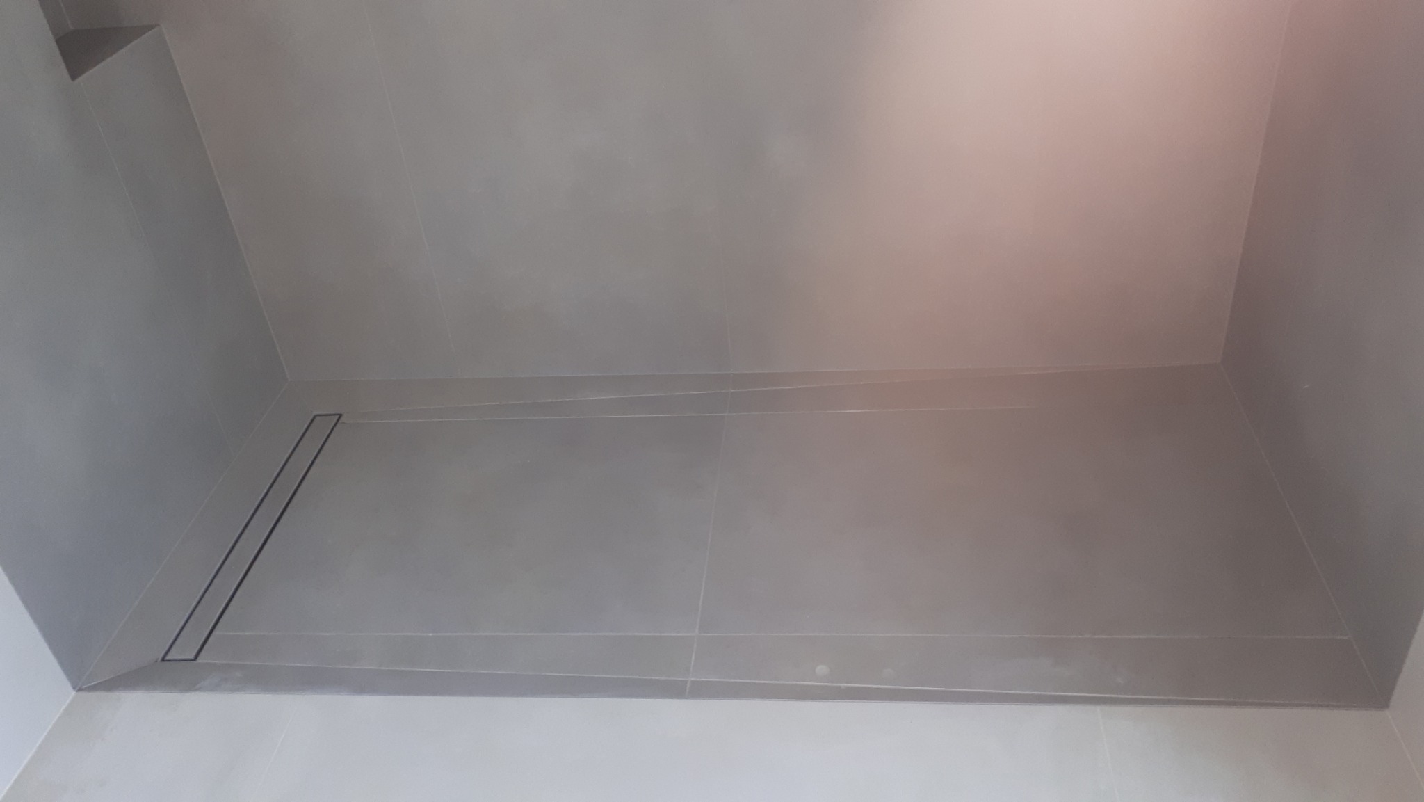 Nowoczesna kabina prysznicowa z odpływem liniowym, wykończona szarymi płytkami. Minimalistyczny design, precyzyjne ułożenie płytek, widoczny fragment półki w rogu.