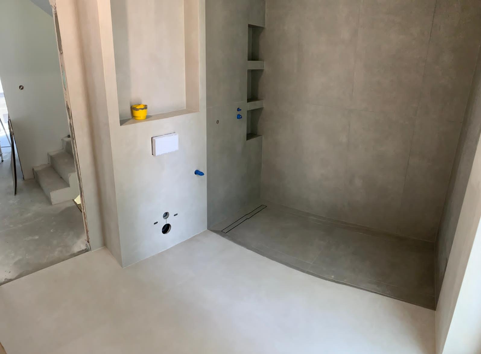 Nowoczesna łazienka w trakcie wykończenia: szare płytki, wnęki w ścianie, biała armatura. Widoczne otwory na instalacje i żółte wiaderko.