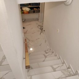 Sonbud - Schody wewnętrzne wyłożone białymi płytkami imitującymi marmur, z widoczną drewnianą poręczą i wejściem do pomieszczenia gospodarczego wykończonego w podobnym stylu.