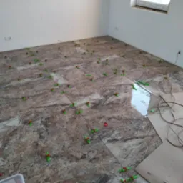 Układanie marmurowych płytek podłogowych z użyciem systemu poziomowania, widoczny pojemnik z czerwonymi klinami i zielone dystanse.