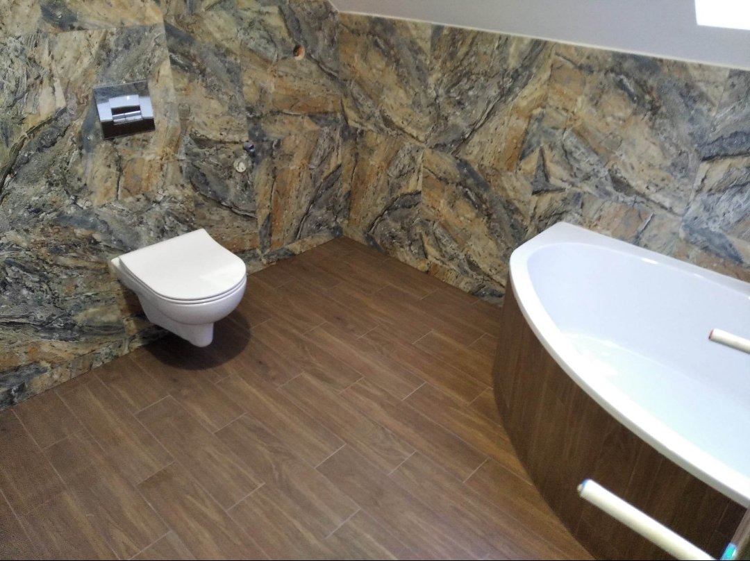 Nowoczesna łazienka z brązowymi płytkami imitującymi drewno na podłodze i ścianach, z elementami dekoracyjnymi przypominającymi kamień, wisząca toaleta i wanna.