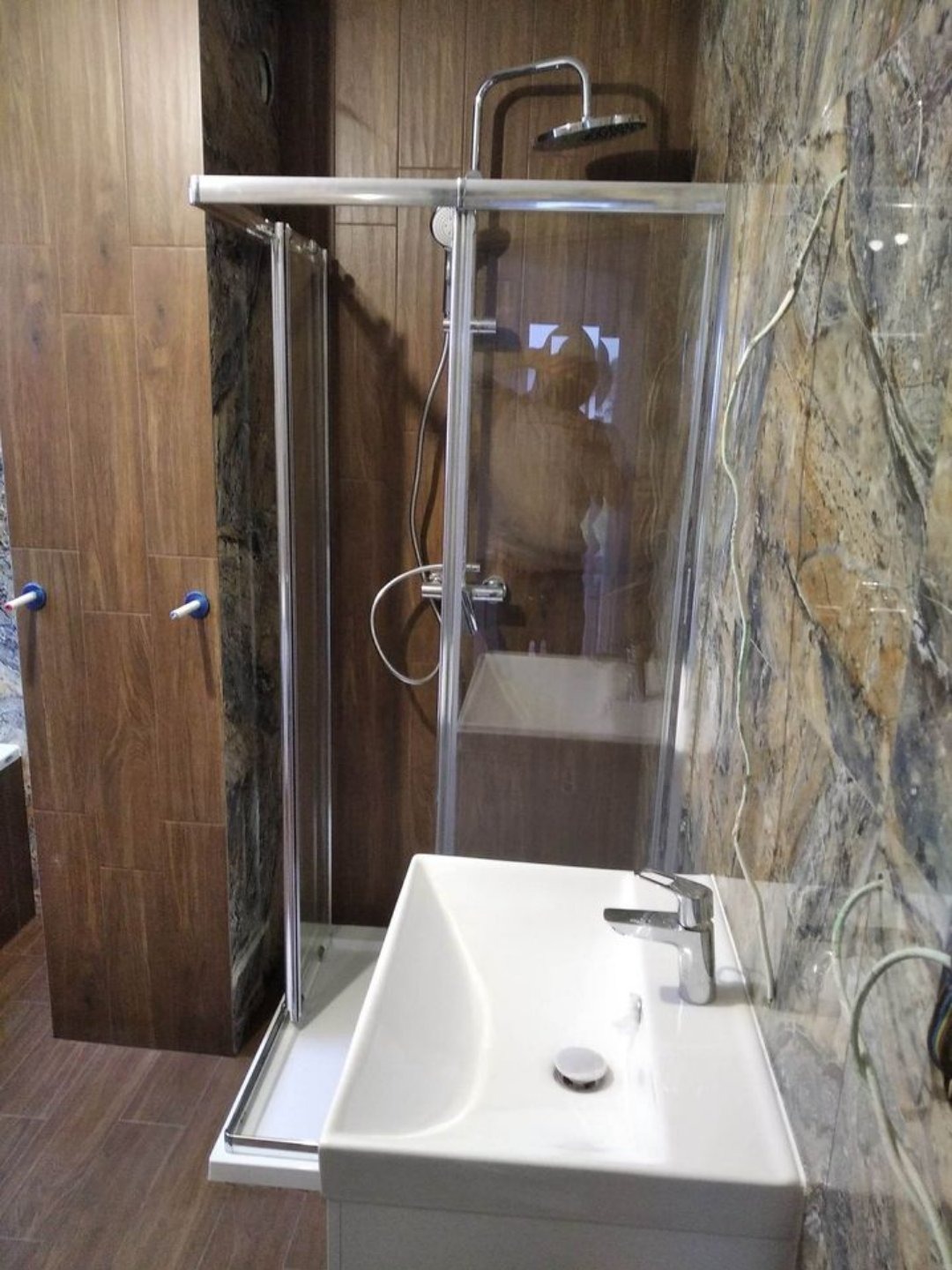 Wykończona łazienka z kabiną prysznicową z przesuwnymi drzwiami, białą umywalką nablatową i brązowymi płytkami imitującymi drewno na ścianach oraz marmurkową mozaiką na jednej ze ścian, widoczne...