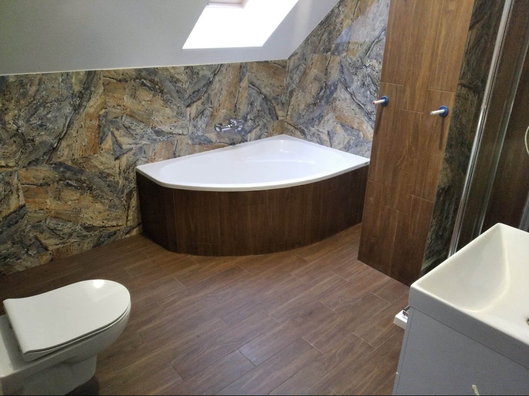 Nowoczesna łazienka na poddaszu z narożną wanną obudowaną drewnopodobnymi panelami, kamienną mozaiką na ścianach, drewnopodobną podłogą i oknem dachowym