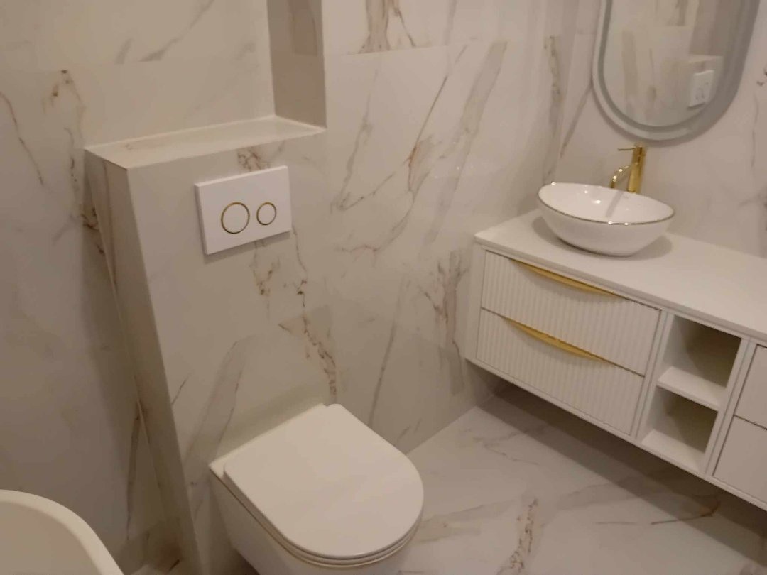 Nowoczesna łazienka wykończona w bieli i marmurze, z wiszącą toaletą, umywalką nablatową ze złotą armaturą i szafką z frezowanymi frontami oraz złotymi uchwytami.