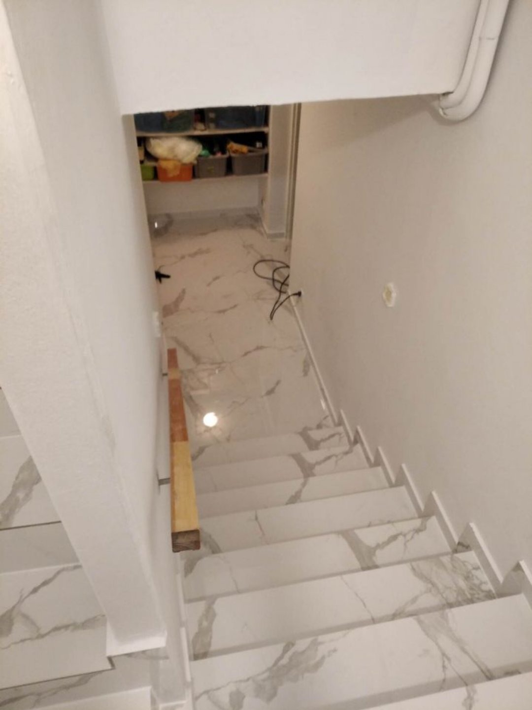 Schody wewnętrzne wyłożone białymi płytkami imitującymi marmur, z widoczną drewnianą poręczą i wejściem do pomieszczenia gospodarczego wykończonego w podobnym stylu.