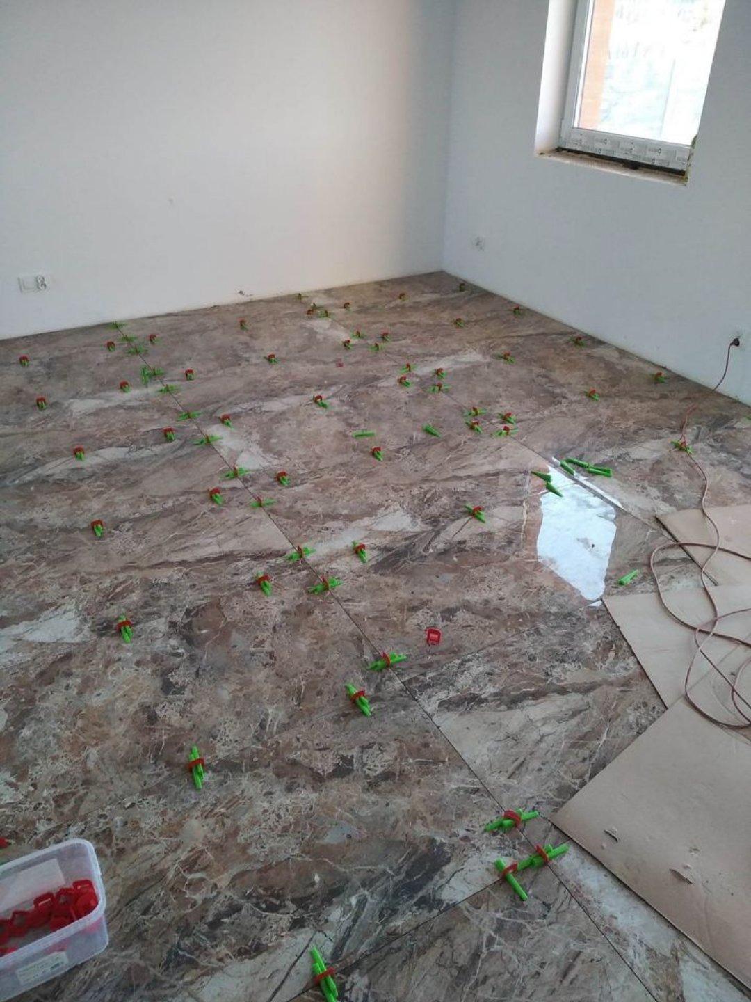 Układanie marmurowych płytek podłogowych z użyciem systemu poziomowania, widoczny pojemnik z czerwonymi klinami i zielone dystanse.