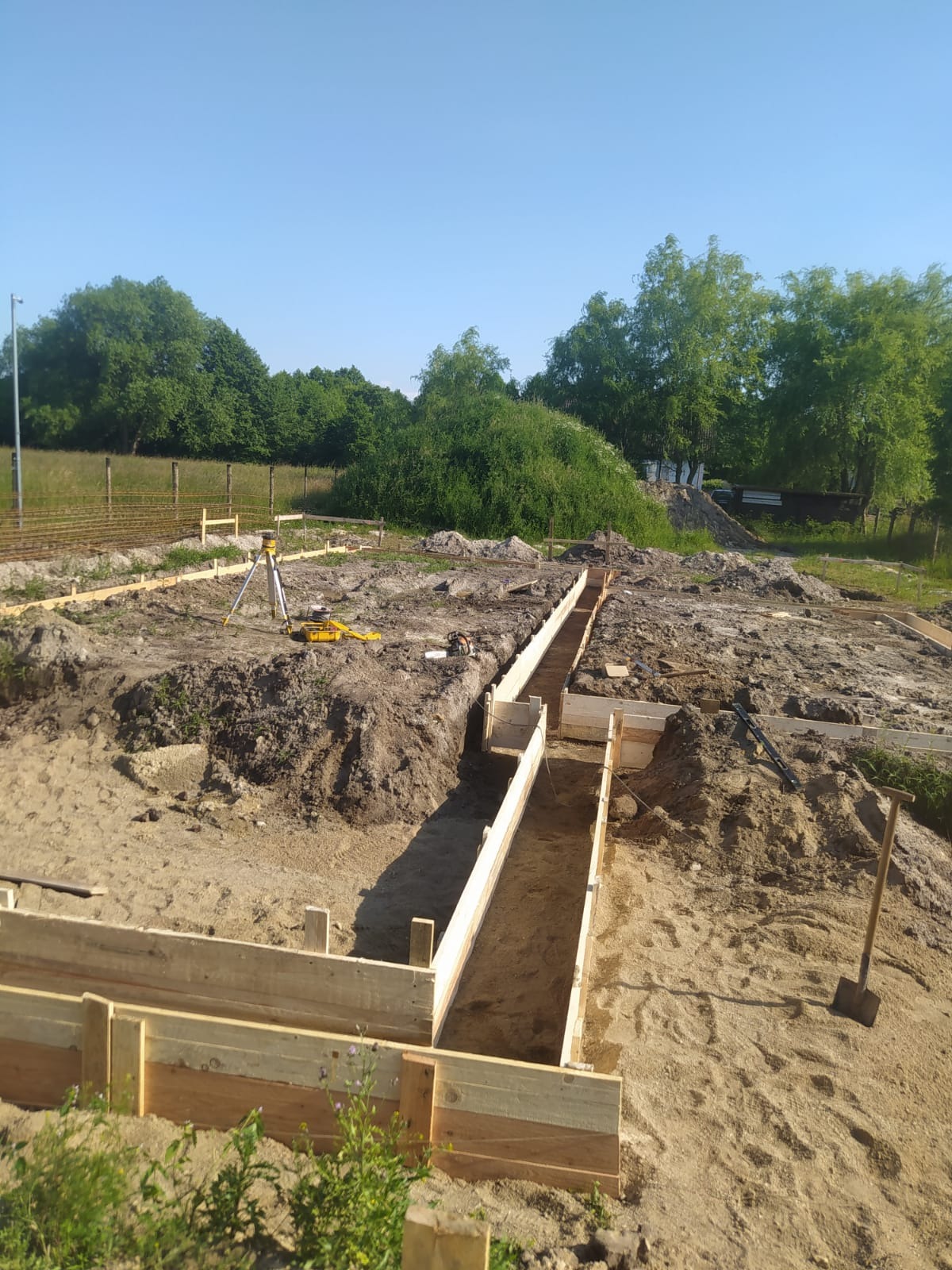 Przygotowanie wykopu pod fundamenty z drewnianym szalunkiem; widoczny niwelator laserowy, teren budowy z ziemią i piaskiem, zieleń w tle.