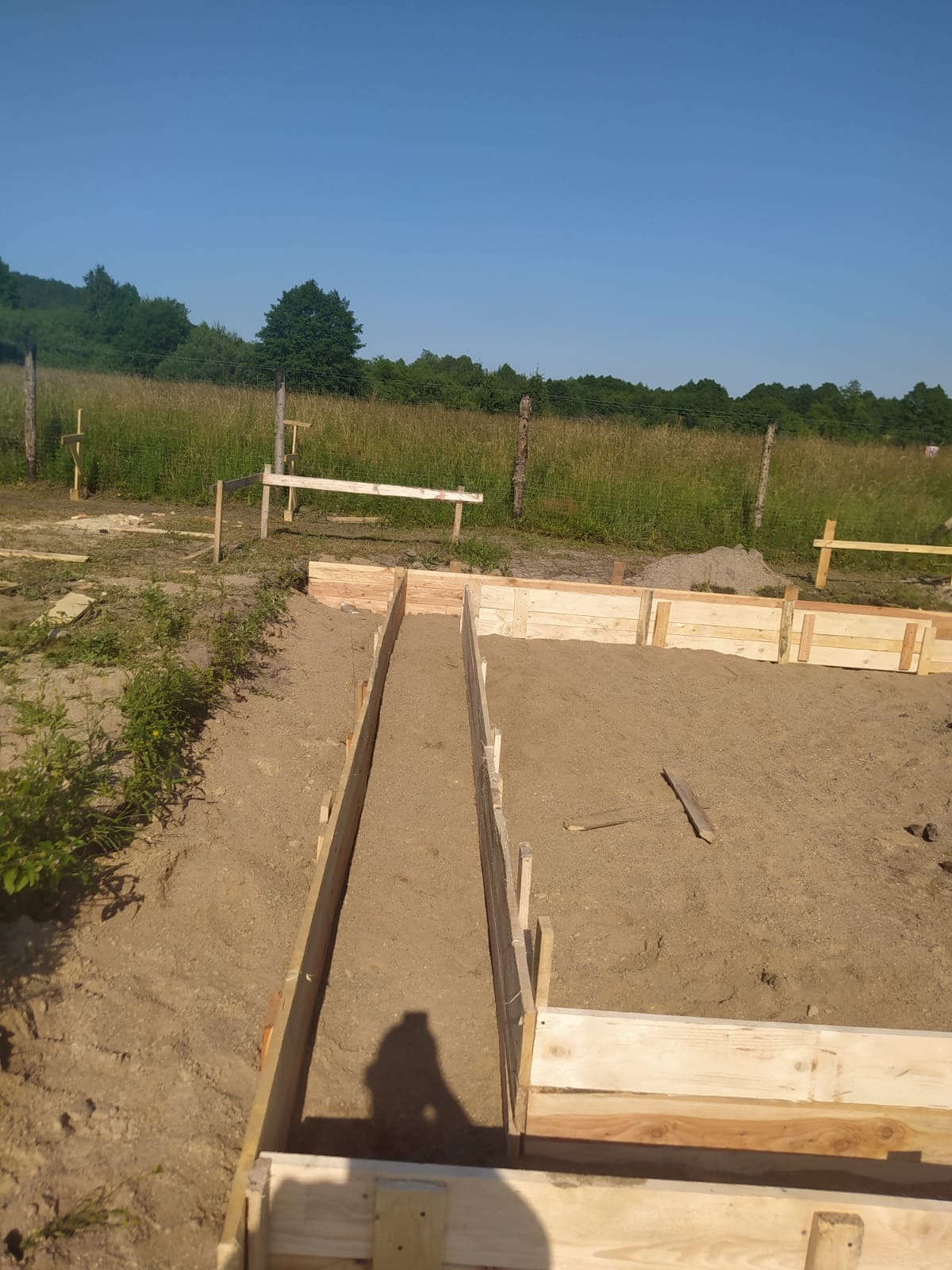 Przygotowanie szalunku pod fundament z wypełnieniem piaskiem, widoczny cień osoby fotografującej, w tle łąka i las.