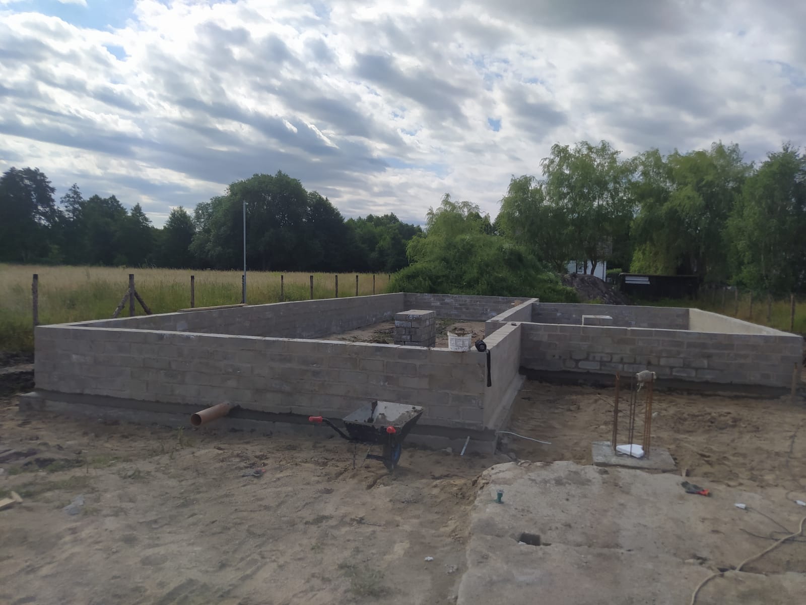 Wzniesione mury fundamentowe z bloczków betonowych na piaszczystym podłożu, widoczna taczka i wystające pręty zbrojeniowe, w tle zieleń drzew i pochmurne niebo.