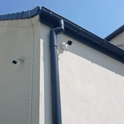 Dwie kamery monitoringu zamontowane na elewacji budynku z grafitowymi rynnami, widok z dołu na błękitne niebo.