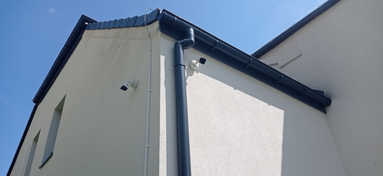 Dwie kamery monitoringu zamontowane na elewacji budynku z grafitowymi rynnami, widok z dołu na błękitne niebo.