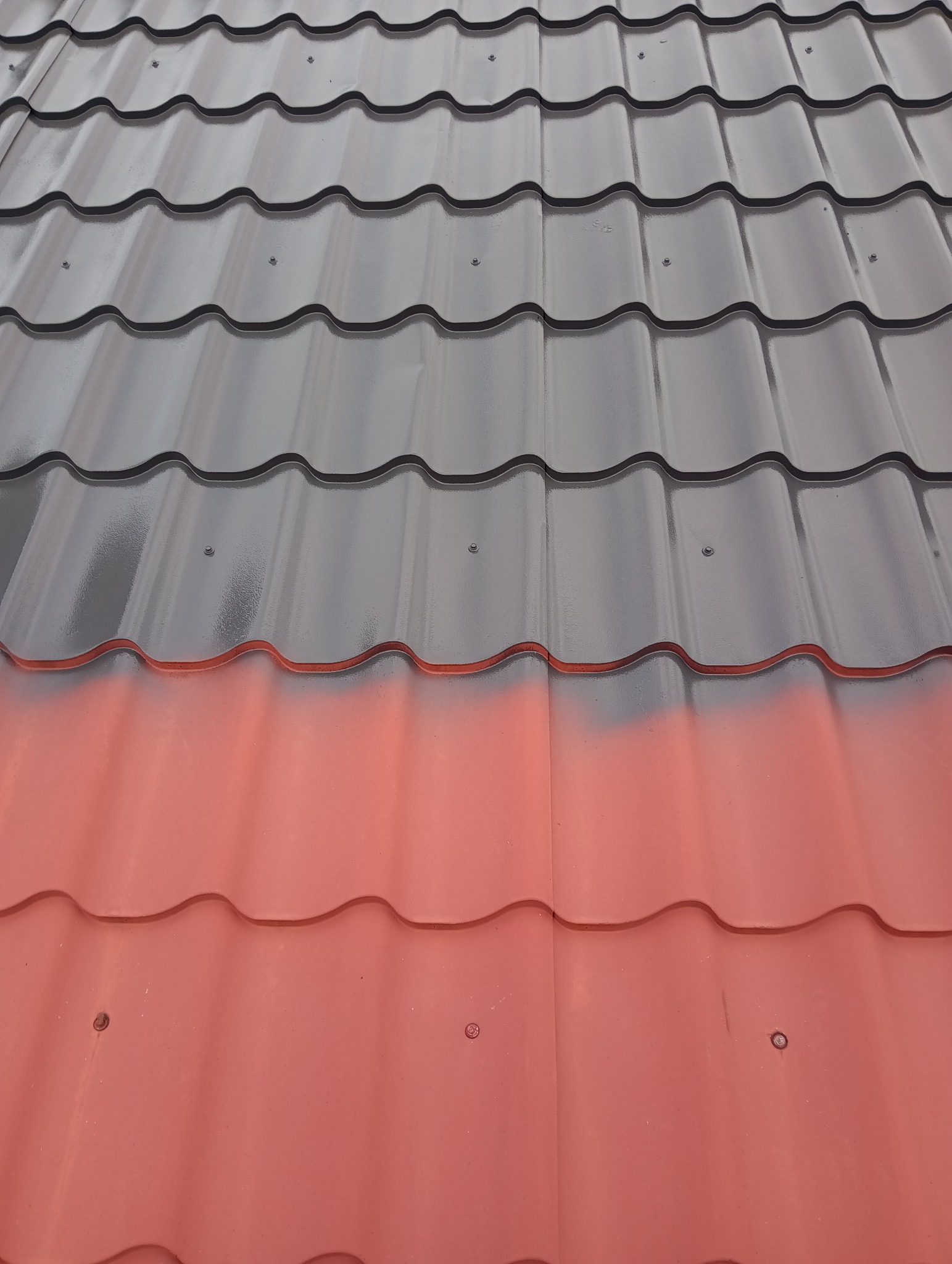 Częściowo pomalowany dach z blachy trapezowej w kolorze grafitowym i ceglastoczerwonym, widoczne mocowania.