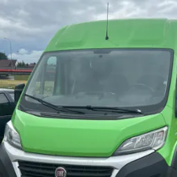 Zielony bus Fiat Ducato z anteną na dachu, widoczny z przodu, z numerem rejestracyjnym KNT 95471, na tle pochmurnego nieba i fragmentu czerwonego mostu.