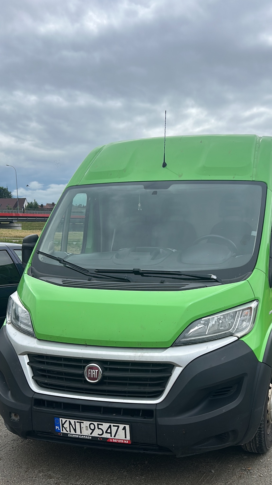Zielony bus Fiat Ducato z anteną na dachu, widoczny z przodu, z numerem rejestracyjnym KNT 95471, na tle pochmurnego nieba i fragmentu czerwonego mostu.