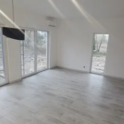 Wnętrze nowo wybudowanego domu z dużymi oknami, szarymi panelami podłogowymi i klimatyzacją na ścianie, widok na plac budowy przez okna.