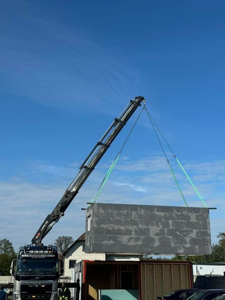 Moduł domu z oknem podnoszony przez dźwig nad kontenerem transportowym na tle błękitnego nieba.