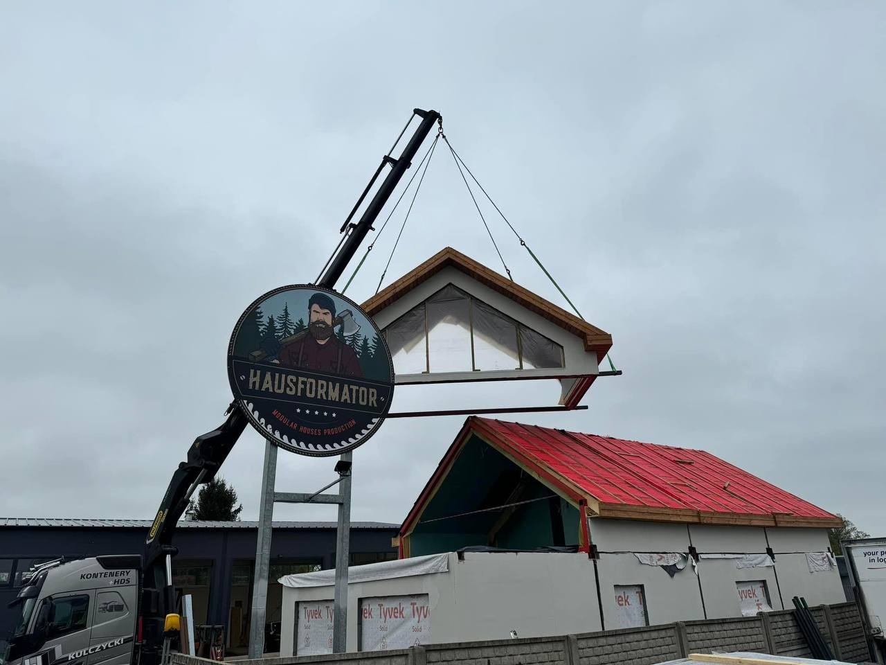 Montaż modułowego segmentu domu przy użyciu dźwigu, widoczne logo firmy Hausformator i fragment budynku z czerwoną membraną dachową.
