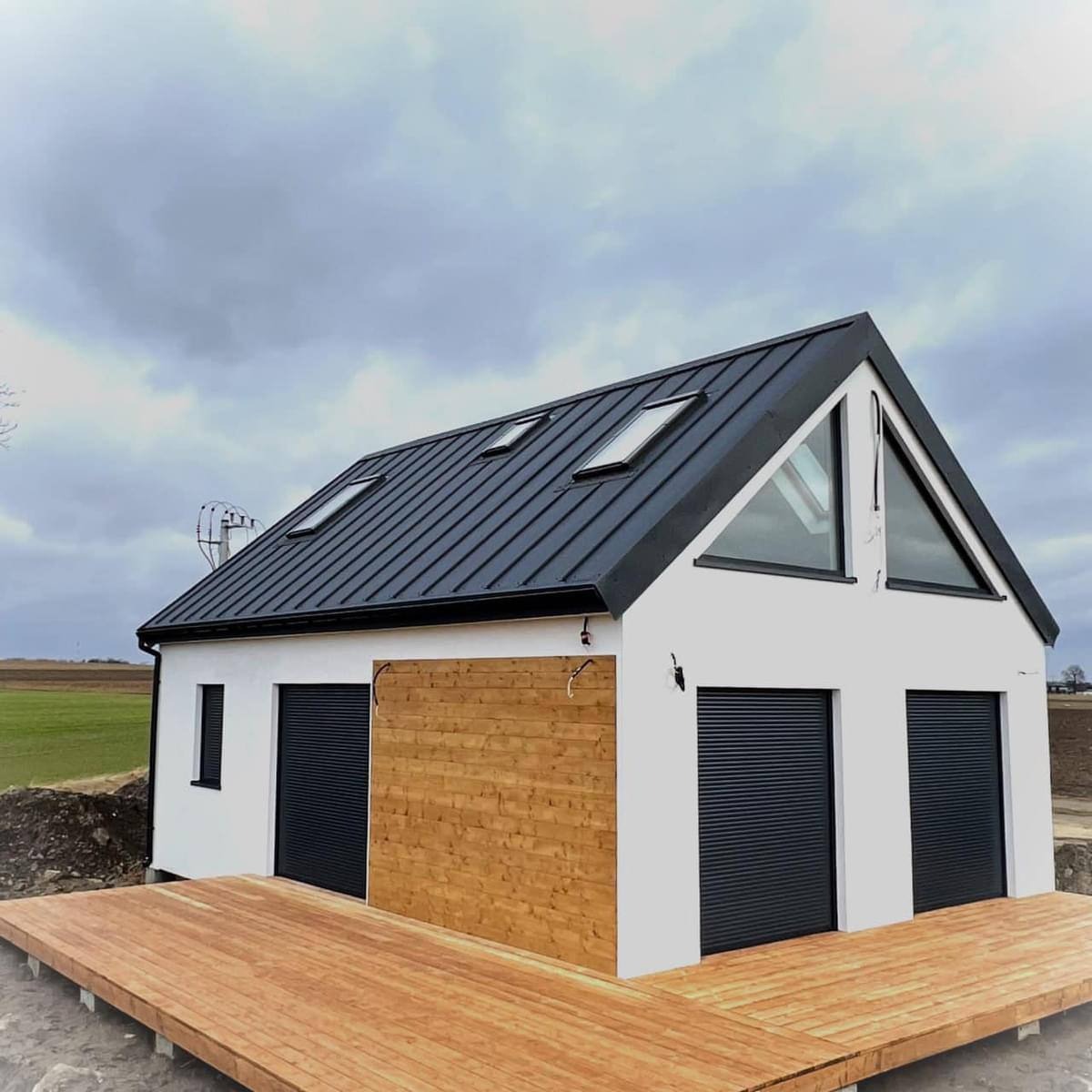 Nowoczesny, biały budynek z czarnym dachem i roletami, drewnianą elewacją oraz tarasem z desek, na tle pochmurnego nieba i zielonego pola.