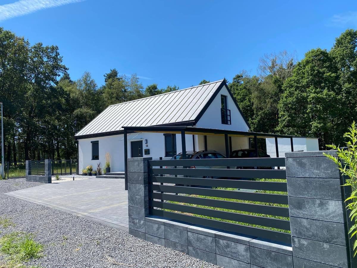 Nowoczesny, parterowy dom z białymi ścianami i metalowym dachem w Bolesławcu, z zadaszonym miejscem parkingowym i szarym ogrodzeniem panelowym.