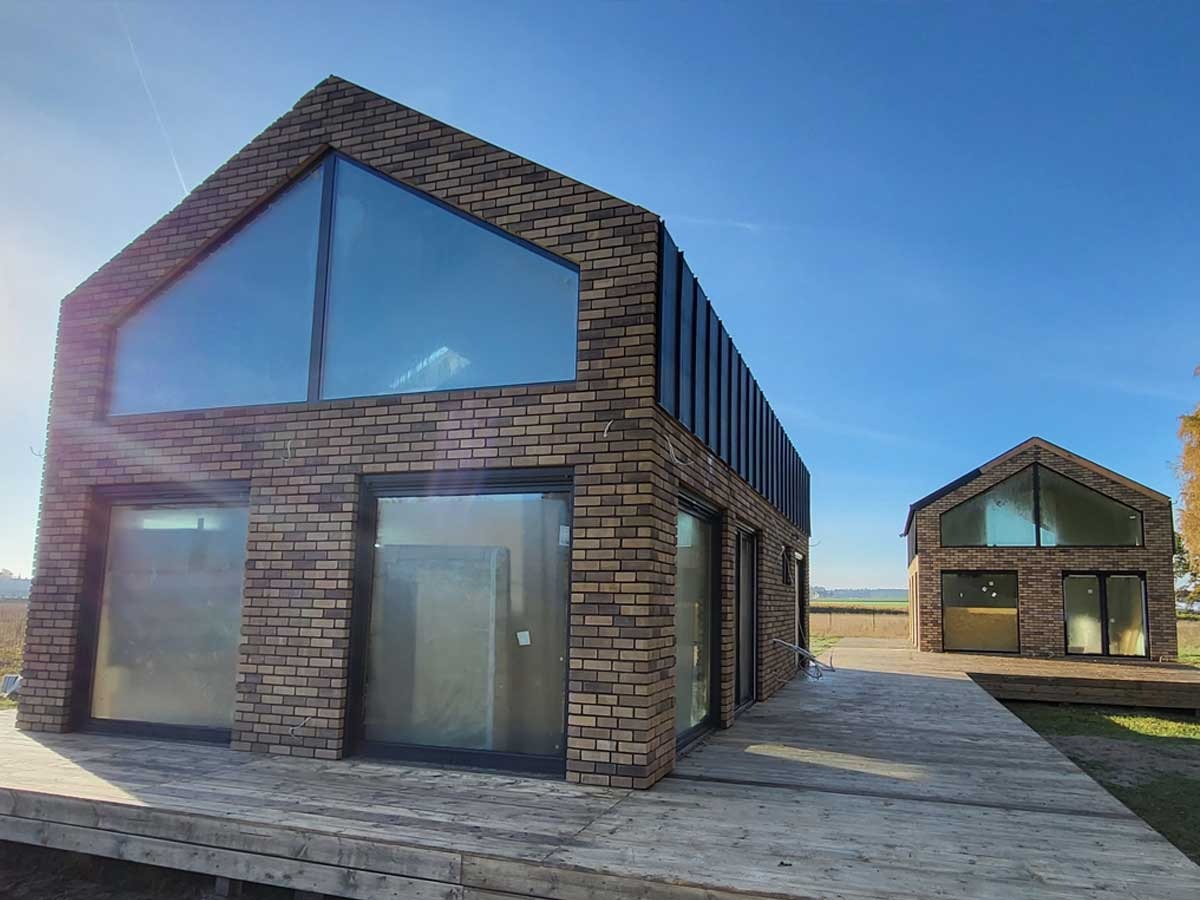 Dwa nowoczesne budynki z cegły klinkierowej z dużymi przeszkleniami, połączone drewnianym tarasem, na tle błękitnego nieba i pól.