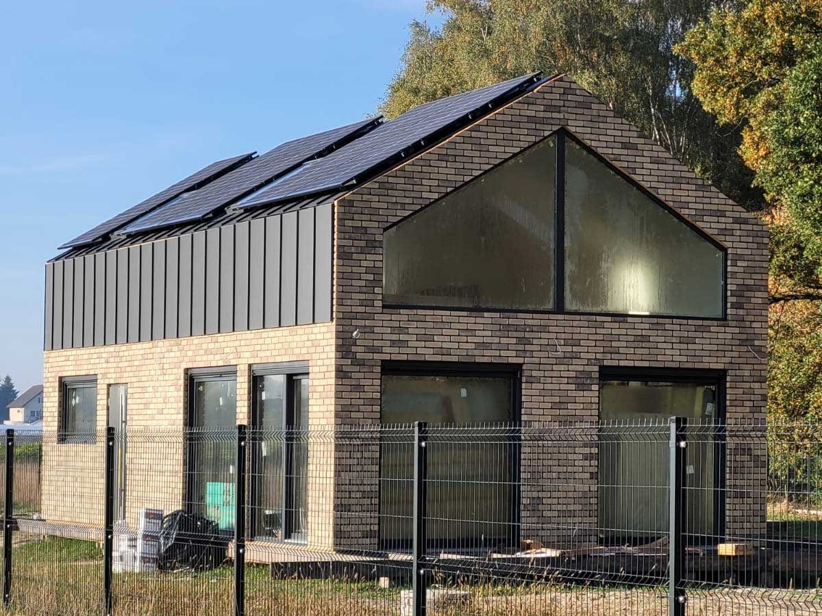 Dwukondygnacyjny dom z cegły klinkierowej z panelami słonecznymi na dachu, widoczne okna z czarnymi ramami i metalowe panele elewacyjne na górnej kondygnacji, ogrodzony siatką.