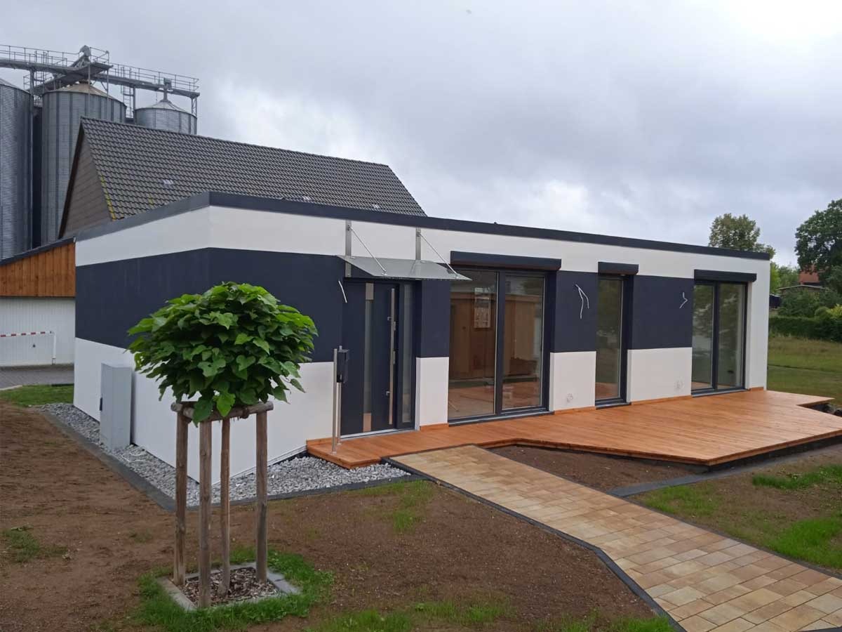 Nowoczesny, parterowy dom z płaskim dachem, wykończony w bieli i granacie, z drewnianym tarasem i chodnikiem z kostki, na tle pochmurnego nieba i silosów zbożowych.