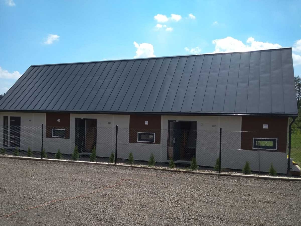 Niski, parterowy budynek z szarą blachą na dachu, wykończony panelami w kolorze białym i brązowym, ogrodzony siatką i obsadzony małymi tujami, na żwirowym podłożu.