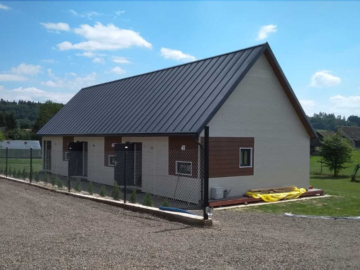 Nowoczesny dom modułowy z ciemnoszarą blachodachówką, jasną elewacją z elementami drewnopodobnymi i czarnym ogrodzeniem siatkowym na żwirowym podłożu, widok z boku w słoneczny dzień.