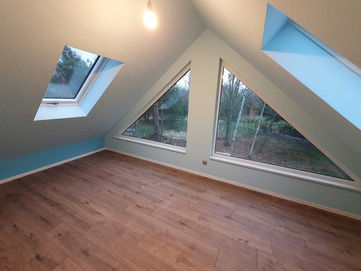 Poddasze z drewnianą podłogą, trójkątnymi oknami z widokiem na drzewa i oknem dachowym, pomalowane na jasny kolor z niebieskim akcentem.