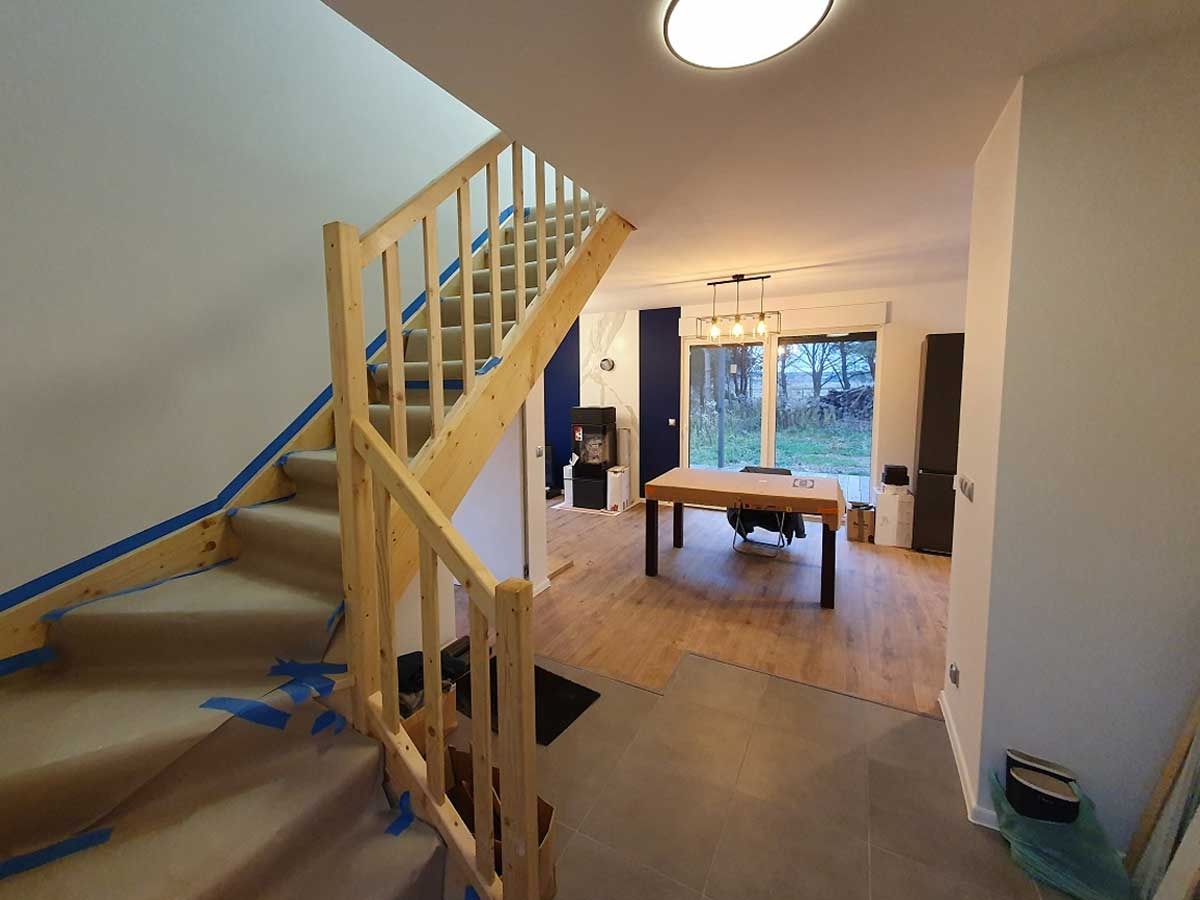 Wnętrze domu w trakcie wykańczania: drewniane schody zabezpieczone taśmą malarską i papierem, widok na salon z oknem tarasowym, fragment położonej podłogi z płytek.