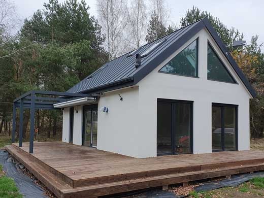 Nowoczesny, biały dom z czarnym dachem i dużymi oknami, z drewnianym tarasem i pergolą, otoczony drzewami w jesiennej scenerii.