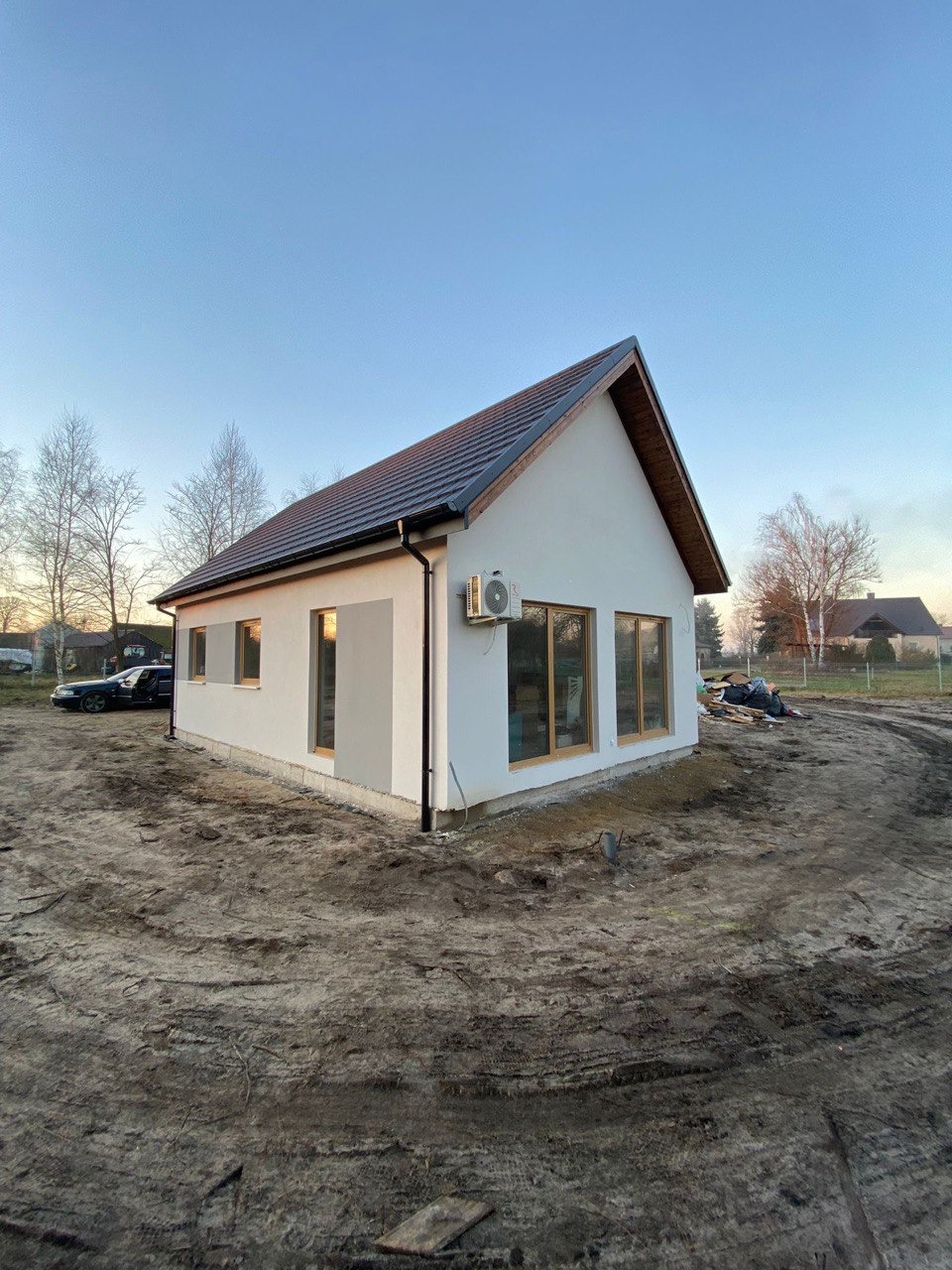 Nieduży, parterowy dom z jasną elewacją i brązowym dachem, z klimatyzatorem na ścianie, na działce z ziemistym podłożem. W tle widoczne drzewa i częściowo zarośnięta działka sąsiednia.