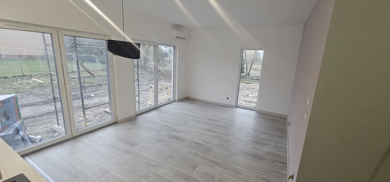 Wnętrze nowo wybudowanego domu z dużymi oknami, szarymi panelami podłogowymi i klimatyzacją na ścianie, widok na plac budowy przez okna.