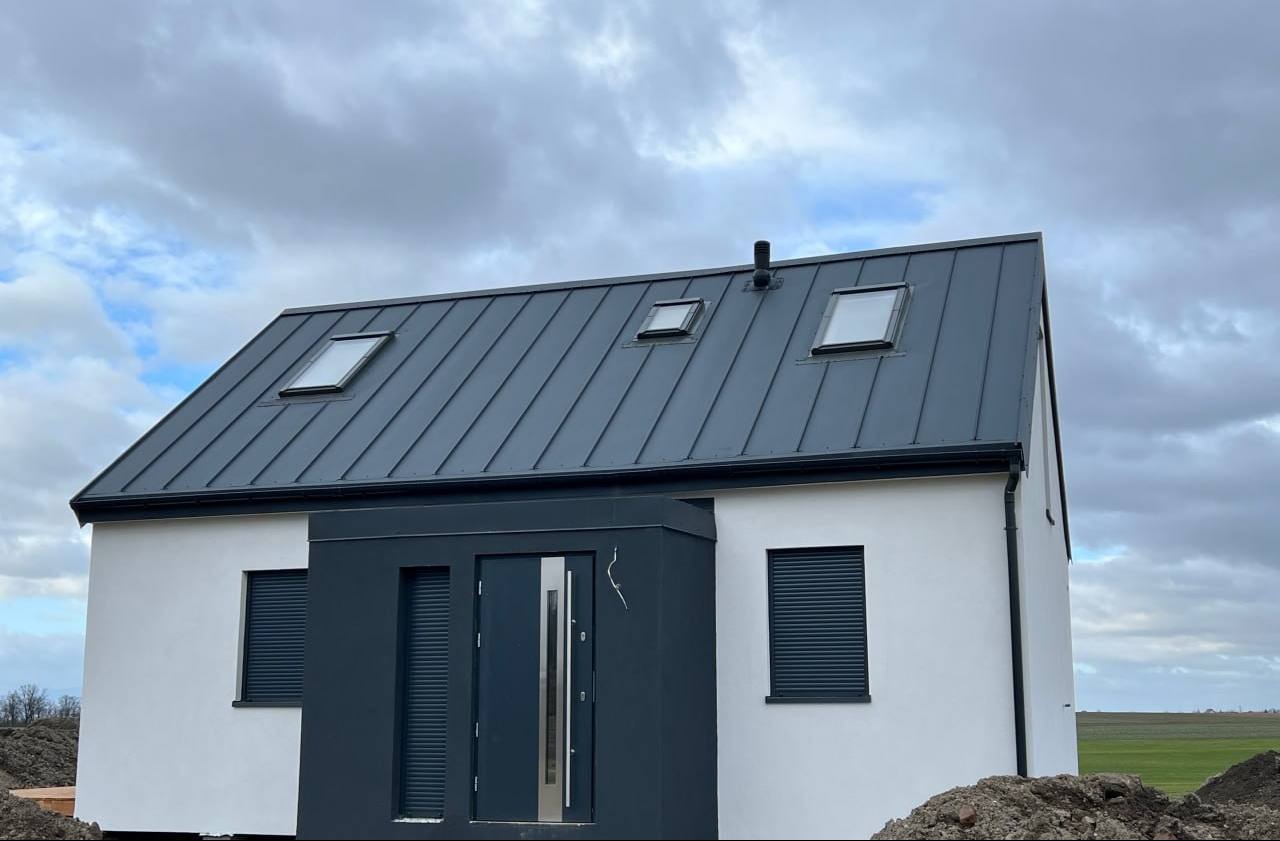 Biały, parterowy budynek z ciemnoszarą, metalową blachodachówką z trzema oknami dachowymi i ciemnoszarym wejściem z roletami zewnętrznymi, na tle pochmurnego nieba.