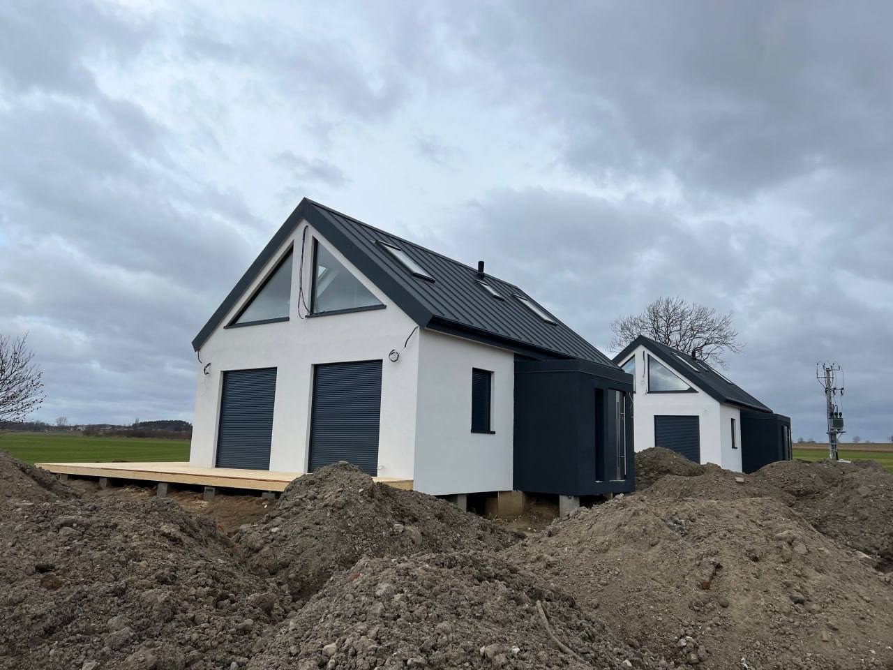 Dwa białe domy modułowe z ciemnymi dachami i roletami, w trakcie budowy, z widocznymi hałdami ziemi i drewnianym tarasem przy jednym z domów, na tle pochmurnego nieba.
